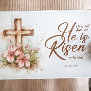 Könnte beinhalten: Ein rechteckiges Glas-Schneidebrett mit einem Aquarelldesign. Es zeigt ein braunes Kreuz, rosa Lilien und den Text "He is Risen" mit dem Bibelvers Matthäus 28,6. Das Gesamtthema ist religiös und hat mit Ostern zu tun.