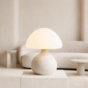 Puede incluir: Una lámpara de mesa con forma de hongo de color crema. La lámpara tiene una base redondeada y una pantalla en forma de cúpula que emite una luz suave y cálida. La lámpara está colocada sobre un soporte blanco en forma de cubo.