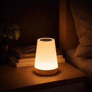 Puede incluir: Una lámpara de noche blanca brillante con una base de madera está sobre una mesita de noche. La lámpara tiene una forma cónica redondeada con una parte superior oscura. Los libros están apilados detrás de la lámpara. La escena está tenue.