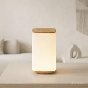 Puede incluir: Lámpara de mesa cilíndrica con tapa y base de madera. La lámpara tiene un cuerpo blanco esmerilado que emite una luz suave y cálida. La lámpara mide aproximadamente 15 cm de alto y está sobre una superficie blanca.