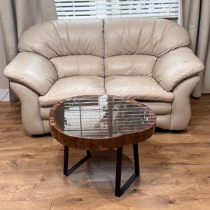 May include: A round coffee table with a dark wood top and black metal legs. The table is in front of a beige leather sofa. The table's top has a natural edge and a glossy finish.