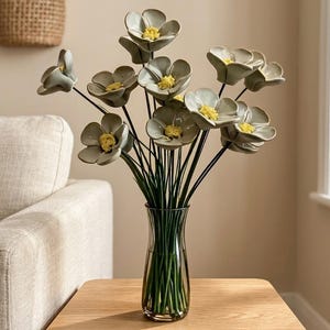 Könnte beinhalten: Ein Strauß aus Keramikblumen mit salbeigrünen Blütenblättern und strukturierten gelben Mitten, angeordnet an dünnen grünen Stielen. Die Blumen stehen in einer klaren, leicht getönten Glasvase mit geschwungener Silhouette und schmalem Hals.