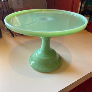 Puede incluir: Un soporte para pasteles de vidrio verde jade vintage. El soporte presenta una plataforma redonda, ligeramente elevada, sostenida por una base de pedestal. El color es un verde suave y opaco, y la pieza tiene un diseño clásico y atemporal. Ideal para exhibir pasteles u otros postres.