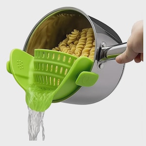 Puede incluir: Un colador de silicona verde para ollas está sujeto a una olla de acero inoxidable llena de pasta. El agua sale del colador. El colador tiene un diseño ranurado para escurrir líquidos.