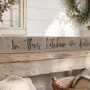 Peut inclure: Une longue enseigne rectangulaire en bois gris patiné portant le texte noir "In this kitchen we dance" écrit en lettres cursives. La surface est rugueuse et texturée, avec un grain de bois visible et des irrégularités naturelles.