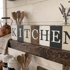 Op de afbeelding: Een rustiek houten bord met het woord "KITCHEN" in grote, vette, witte letters. Het bord is verdeeld in afwisselende secties van wit, bruin en zwart. Het bord wordt weergegeven op een donkere houten plank, met keukengerei en decor in de achtergrond.