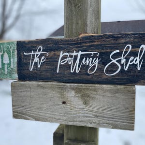May include: A weathered wooden sign, reading "the potting shed" in white script. The sign is made of dark stained wood and features a green section with gardening tools. It is mounted on a wooden post.