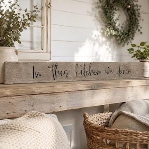 Op de afbeelding: Een lang, rechthoekig houten bord met een verweerde grijze afwerking. De tekst "In This Kitchen We Dance" is in zwarte cursieve letters in het midden van het bord gedrukt. Het bord heeft een ruwe, natuurlijke houtnerfstructuur.