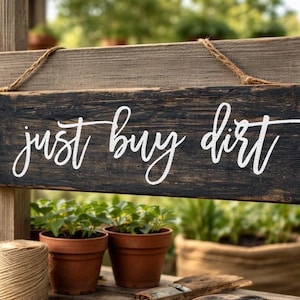 May include: A rectangular wooden sign with a dark, weathered finish hanging from a jute rope. The sign features the white cursive text "just buy dirt" and a green section on the left side displaying white line art of a garden fork, trowel, and rake.