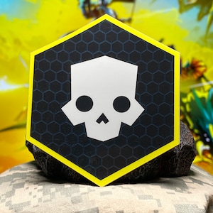 May include: A white skull sticker with black eyes on a black hexagonal background with a yellow border. The sticker is on a rock.