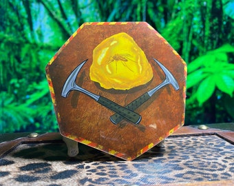 May include: A sticker featuring a yellow amber stone with a fossilized insect inside. Two crossed pickaxes are behind the stone. The sticker has a brown background with a yellow and red border.