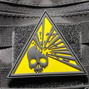 Puede incluir: Un parche negro y amarillo con un diseño de calavera y explosión. La calavera es negra, sobre un fondo amarillo. El parche triangular tiene un borde negro. Está adherido a una tela negra.