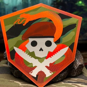 Peut inclure: Un crâne rouge, blanc et noir avec un serpent rouge enroulé autour de lui. Le serpent a un couteau blanc dans sa bouche. Le crâne porte un béret rouge. L'arrière-plan est un motif de camouflage vert et noir.