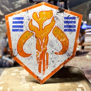 May include: A weathered, hexagonal sign featuring an orange and yellow skull emblem with blue stripes on a white background. The sign has an orange border and a distressed, vintage appearance, suggesting a collectible or fan item.