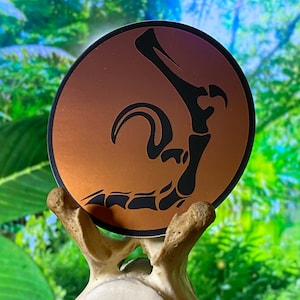 May include: A round sticker with a copper gradient background and a black silhouette of a dinosaur foot.