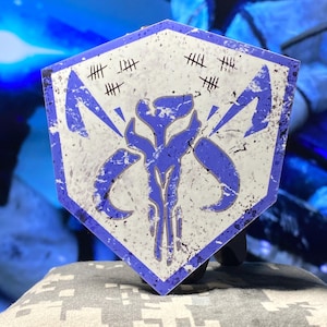 May include: A hexagonal badge featuring a blue and white design with a distressed, weathered appearance. The central image is a stylized blue emblem, surrounded by a white border and a series of tally marks. The badge is likely a collectible or emblem.