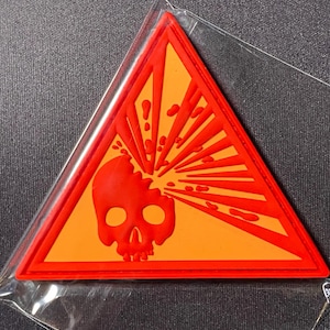 May include: A red and orange patch in a triangular shape, featuring a skull with an explosion graphic. The patch is enclosed in a clear plastic wrapper. The design is a warning symbol.