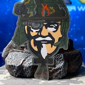 May include: A sticker featuring a cartoon illustration of a soldier wearing a camouflage helmet and eye patch. The soldier has a stern expression and a flame emblem on the helmet. The sticker is placed on a textured surface.