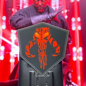 May include: A black and red Mandalorian symbol sticker on a black background. The sticker is shaped like a shield and has a red skull with horns and stripes.