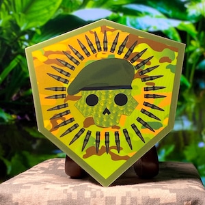 May include: A hexagonal-shaped military-themed emblem featuring a skull wearing a green beret, surrounded by bullets. The background has a camouflage pattern in shades of green and yellow. The emblem is framed with a green border.
