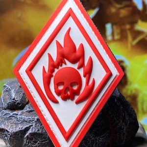 Pode incluir: Um remendo em forma de diamante vermelho e branco com uma caveira e chamas no centro. A caveira é branca e as chamas são vermelhas.