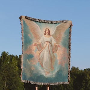 May include: A woven throw blanket featuring a classical painting of an angel with large wings, set against a blue and pink sky. The blanket has a fringed edge and the text "The Sanctum Atelier" at the top.