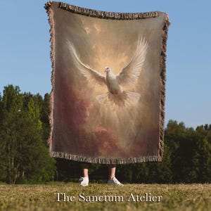 May include: A woven blanket featuring a white dove with outstretched wings against a backdrop of soft, warm colors. The blanket has a fringed edge and the text "The Sanctum Atelier" at the bottom. The blanket is held up against a blue sky.