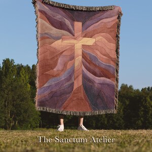 May include: A woven throw blanket featuring a cross design in shades of brown, purple, and tan. The blanket has a fringed edge and is held up against a blue sky. The text "The Sanctum Atelier" is at the bottom.