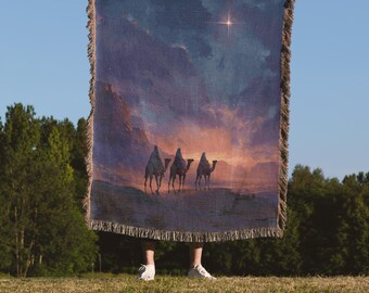 Op de afbeelding: Een rechthoekige blauwe geweven sprei met een landschapsscène van drie figuren op kamelen, een heldere ster en een zonsondergang. De sprei heeft een gefranjerde rand en de tekst "The Sanctum Atelier" onderaan.