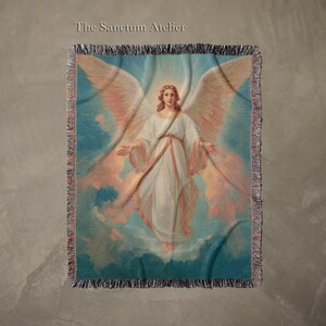 May include: A woven throw blanket featuring a celestial image of an angel with large wings, dressed in white robes, against a backdrop of clouds and a blue sky. The text "The Sanctum Atelier" is at the top. The blanket has a fringed edge.