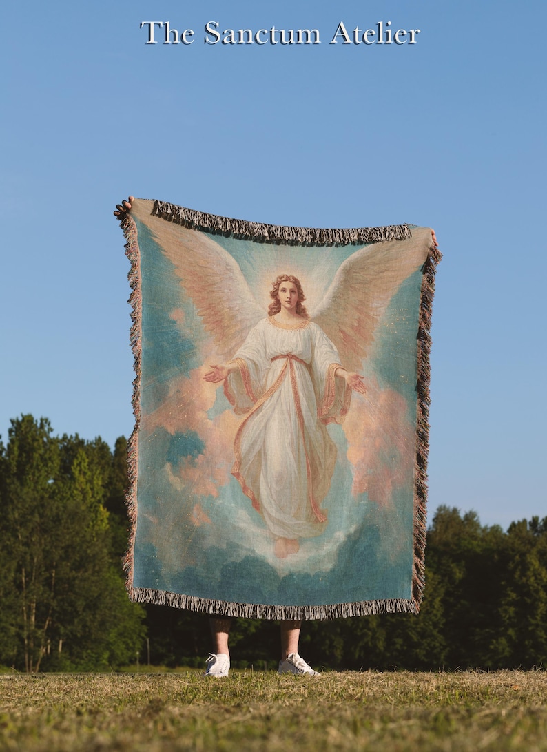 May include: A woven throw blanket featuring a classical painting of an angel with large wings, set against a blue and pink sky. The blanket has a fringed edge and the text "The Sanctum Atelier" at the top.
