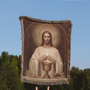 May include: A woven throw blanket featuring a religious depiction of Jesus holding a communion host above a chalice, with wheat and grapes on a table. The design includes a halo and arched background. The text TheSanctumAtelier appears at the top.