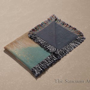 May include: Folded throw blanket with a teal and tan watercolor design, accented with gold flecks. The blanket has a dark gray side and is finished with a fringe trim. The Sanctum Atelier logo is in the bottom right corner.