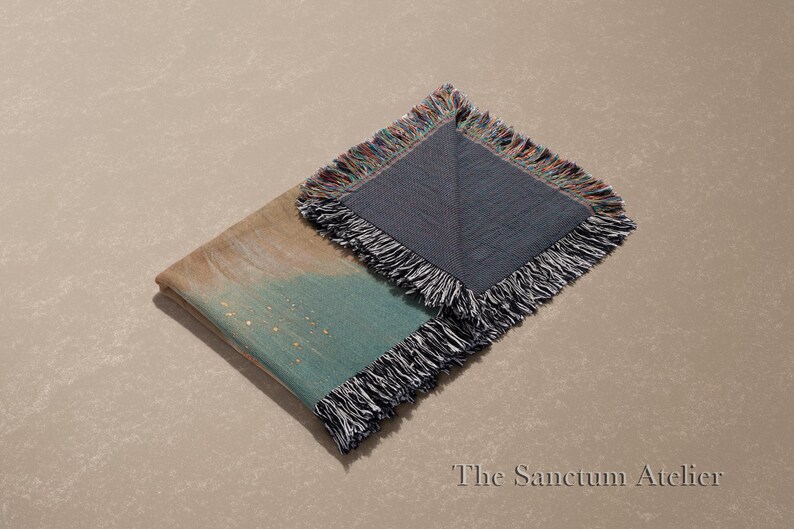 May include: Folded throw blanket with a teal and tan watercolor design, accented with gold flecks. The blanket has a dark gray side and is finished with a fringe trim. The Sanctum Atelier logo is in the bottom right corner.
