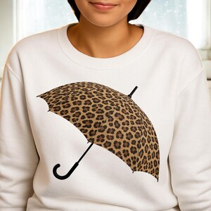Peut inclure: Sweat-shirt blanc avec un motif de parapluie à imprimé léopard. Le parapluie a une poignée et une armature noires, et l'imprimé léopard est marron et beige. Le sweat-shirt est de style décontracté.