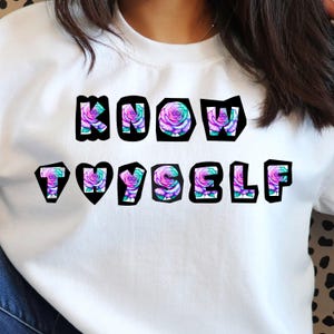 Può includere: Felpa bianca con la scritta "KNOW YOURSELF" in lettere nere bordate. Ogni lettera è riempita con un disegno di rosa ad acquerello nei toni del viola, verde acqua e verde. La felpa è a girocollo.