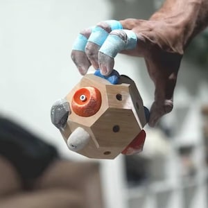 Peut inclure: Prise d'escalade en bois avec différentes prises colorées, notamment orange, grise et bleue. La prise est tenue par une main avec du ruban adhésif bleu sur les doigts. La prise a une forme géométrique et est conçue pour l'escalade.