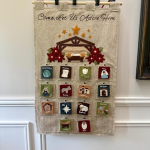 May include: A beige felt Advent calendar with the text "Come, Let Us Adore Him." It features a nativity scene with a star and red flowers. Below are 16 small squares with religious illustrations and text.