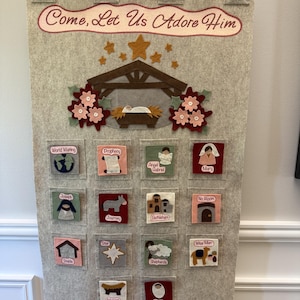 Könnte beinhalten: Ein beigefarbener Wandbehang aus Filz mit dem Text Come, Let Us Adore Him, einer Krippenszene und sechzehn quadratischen Taschen. Jede Tasche zeigt eine Filzapplikation zu einem weihnachtlichen Thema, darunter Maria, Josef und das Jesuskind.
