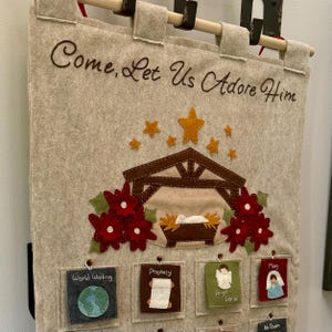 May include: Felt Christmas advent calendar with the words "Come, Let Us Adore Him." The calendar features a nativity scene with a star, flowers, and pockets with images and text, including "World Waiting," "Prophecy," and "Angel Gabriel."