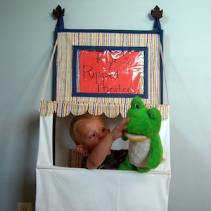 Puede incluir: Un niño está usando un teatro de títeres hecho en casa con un títere de rana verde. El teatro está hecho de tela y tiene un letrero que dice "Rye's Puppet Theater".