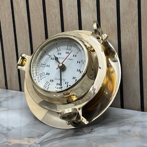 May include: A polished brass porthole clock with a white face, featuring Roman numerals for 1 through 12 and smaller Arabic numerals for 13 through 23. The clock face includes the text "SHIP TIME" and "QUARTZ" with black hour, minute, and second hands.