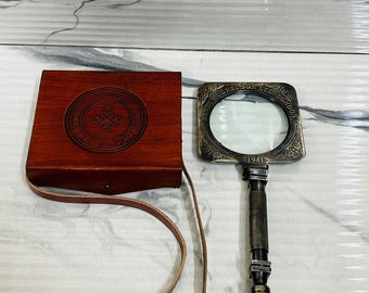 May include: A vintage-style magnifying glass with a square lens and ornate metal frame, next to a wooden box with a leather strap. The magnifying glass has the text "HENRY HUGHES & SON LTD LONDON 1941".