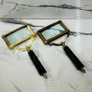 May include: Two antique-style magnifying glasses with rectangular lenses and ornate brass frames. The handles are black with carved detailing and gold-coloured accents. The magnifying glasses are displayed on a marble-patterned surface.