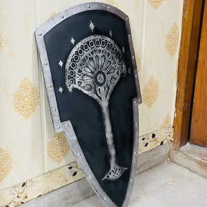 Peut inclure: Un bouclier en forme de cerf-volant avec une finition noire mate et une bordure argentée métallique ornée de rivets visibles. Le centre présente un emblème argenté en relief représentant un arbre avec une canopée en éventail.
