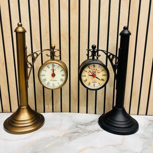 Può includere: Due orologi decorativi a doppia faccia. Uno è dorato con un quadrante color crema e numeri romani. L'altro è nero con un quadrante beige e numeri romani. Entrambi hanno un'estetica vintage.