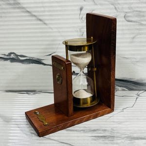Peut inclure: Un sablier en bois avec des détails en laiton, placé dans un cadre en bois foncé. Le sablier a des ampoules en verre transparent remplies de sable blanc. Le cadre comporte du matériel décoratif en laiton. Un dispositif de chronométrage classique.
