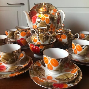 Pode incluir: Um conjunto de chá completo com bule, açucareiro, cremeira e várias chávenas e pires. O conjunto apresenta um design pintado à mão de fruta laranja e folhas verdes sobre um fundo branco com detalhes dourados. O conjunto é exibido numa superfície de madeira.