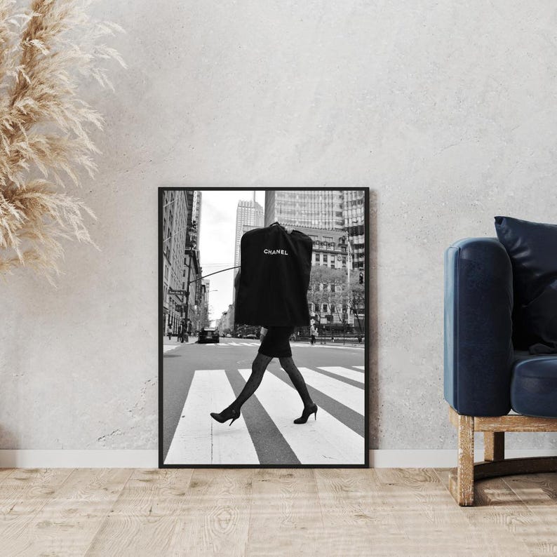 May include: Black and white framed photograph of a person crossing a city street in high heels. The person is wearing a black dress and carrying a garment bag with the Chanel logo. The background features buildings and a crosswalk.