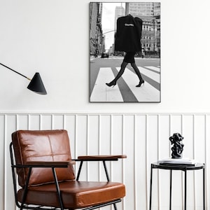 May include: Black and white framed photograph of a person crossing a crosswalk in a city. The person is wearing a black dress and heels, carrying a garment bag with the word "CHANEL" printed on it. The background features buildings and a street.